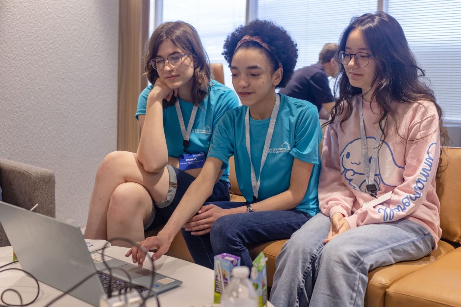 Tres mujeres jóvenes sentadas en un sofá, enfocadas en una computadora portátil. Dos usan camisas y cordones azules a juego, mientras que el tercero usa una sudadera rosa. El estado de ánimo es colaborativo.
