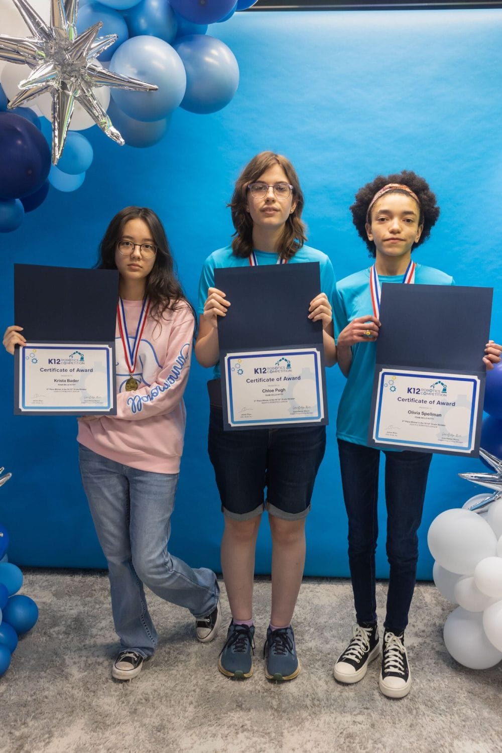 Tres estudiantes están uno al lado del otro sosteniendo certificados, con medallas. Están rodeados de globos azules y blancos, que exudan un ambiente de celebración.