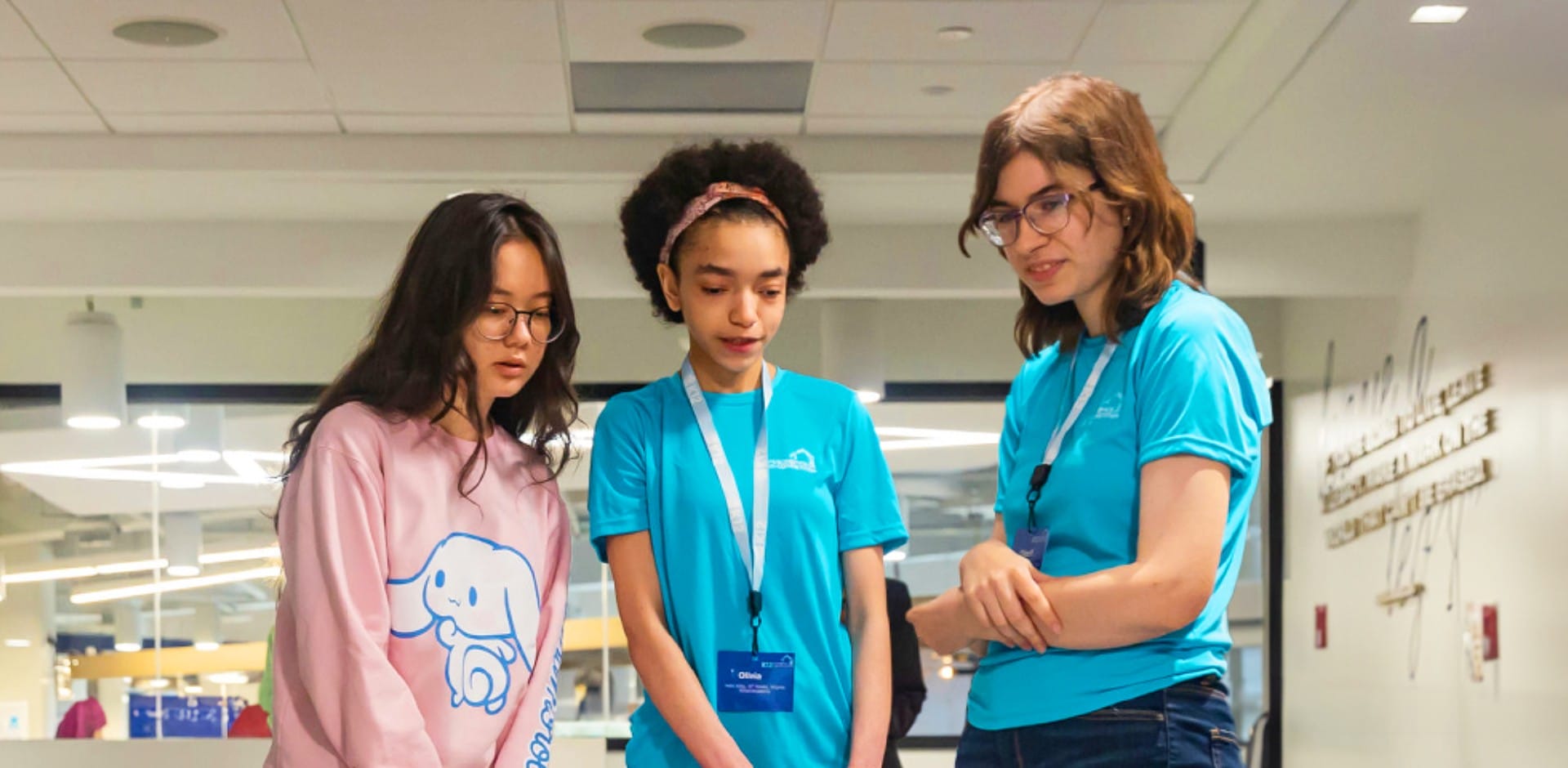 Three girls wearing blue shirts stand together in a hallway, smiling and engaging with one another.