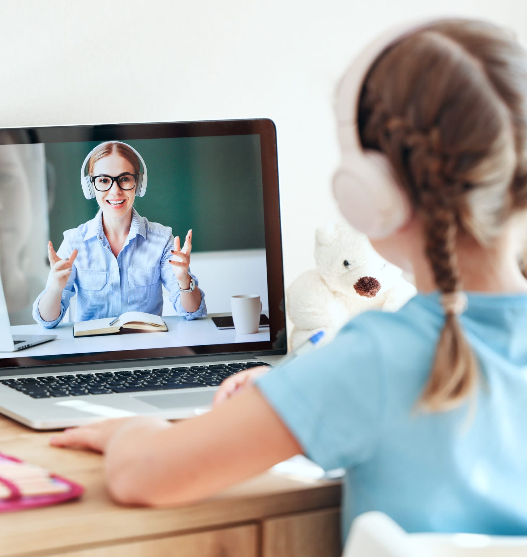 A young girl sits at a desk wearing headphones, focused on a laptop displaying a woman on the screen.