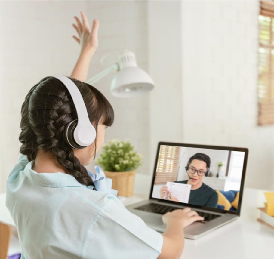 A girl wearing headphones raises her hand while participating in an online class on a laptop.