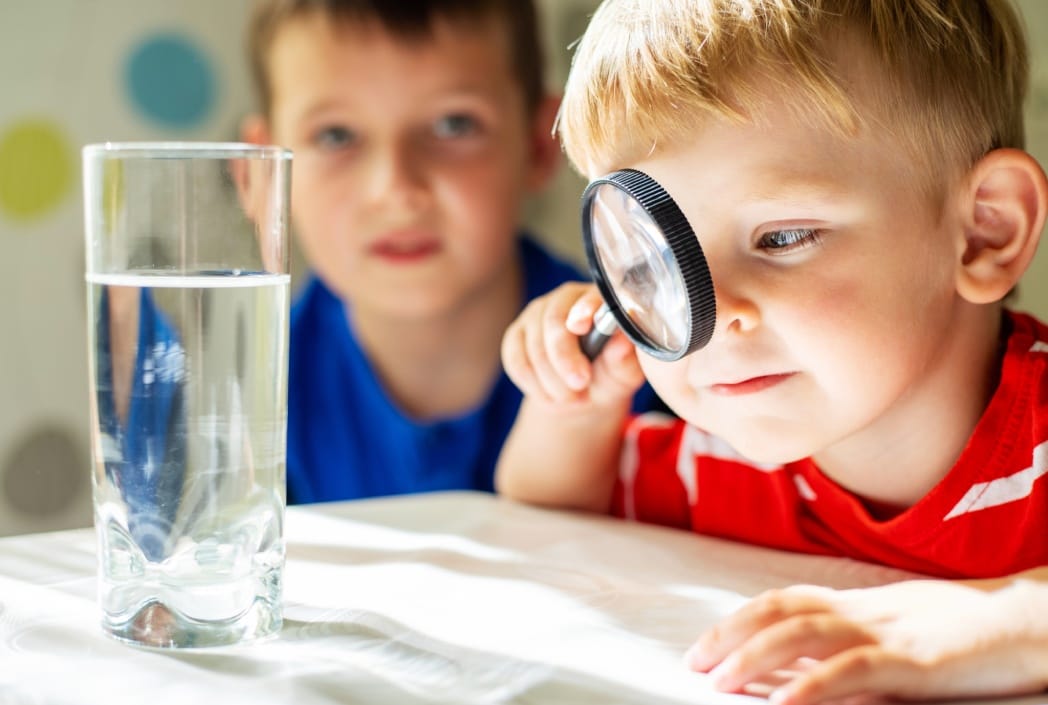 Un niño con camisa roja examina un vaso de agua con una lupa, mientras que otro niño con camisa azul observa. Ambiente curioso y lúdico.