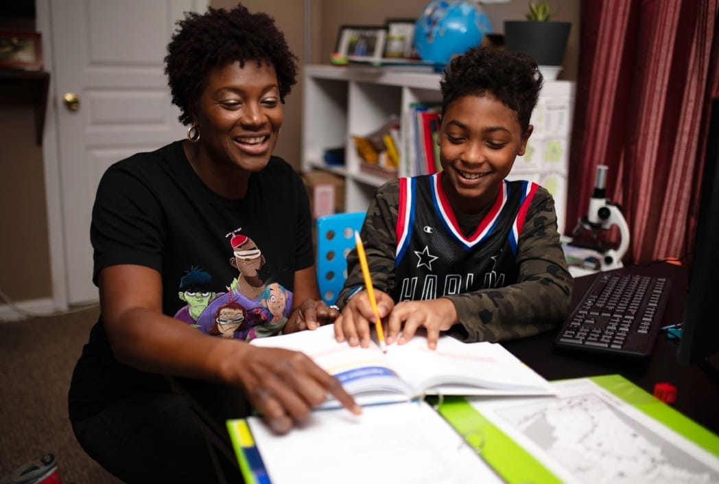 Una mujer y un niño sonrientes trabajan juntos en la tarea en un escritorio. El niño sostiene un lápiz, mientras que la mujer señala un libro de texto, creando una atmósfera de aprendizaje alegre y colaborativa.