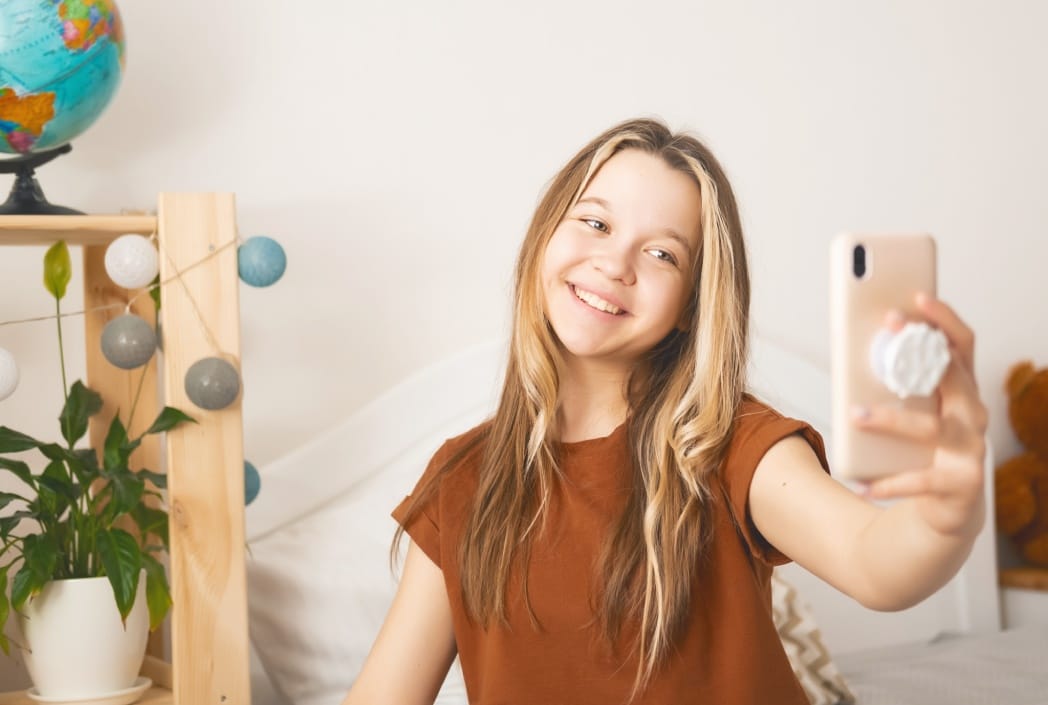 A smiling girl in a brown shirt takes a selfie in a cozy room. A globe and a plant with string lights decorate the background, creating a cheerful mood.