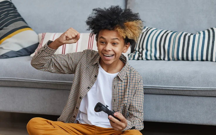 A young person sitting on a couch, holding a game controller, smiles joyfully with a fist raised. The background has striped cushions, creating a casual and happy atmosphere.