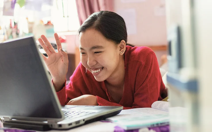 Smiling woman in a red sweater waves at a laptop screen, conveying a joyful interaction. She's lying on a bed with a book nearby in a cozy room.