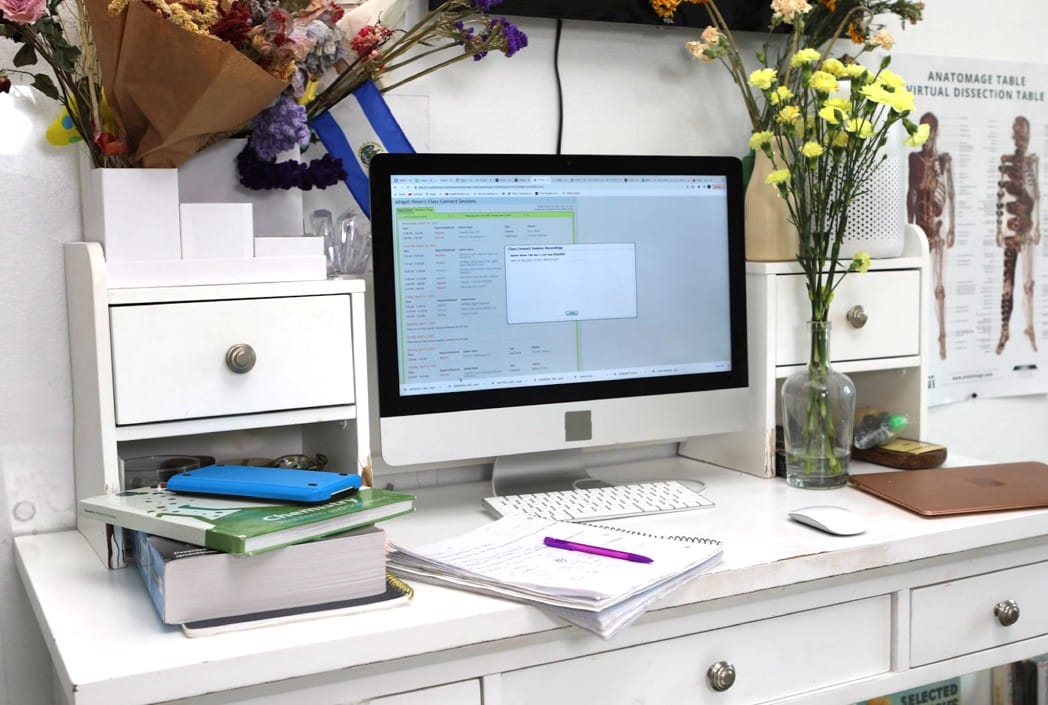 Un escritorio blanco desordenado con un monitor de computadora que muestra una pantalla de datos. Libros, notas y un bolígrafo yacen cerca. Las flores coloridas están en jarrones, agregando un toque vivo.