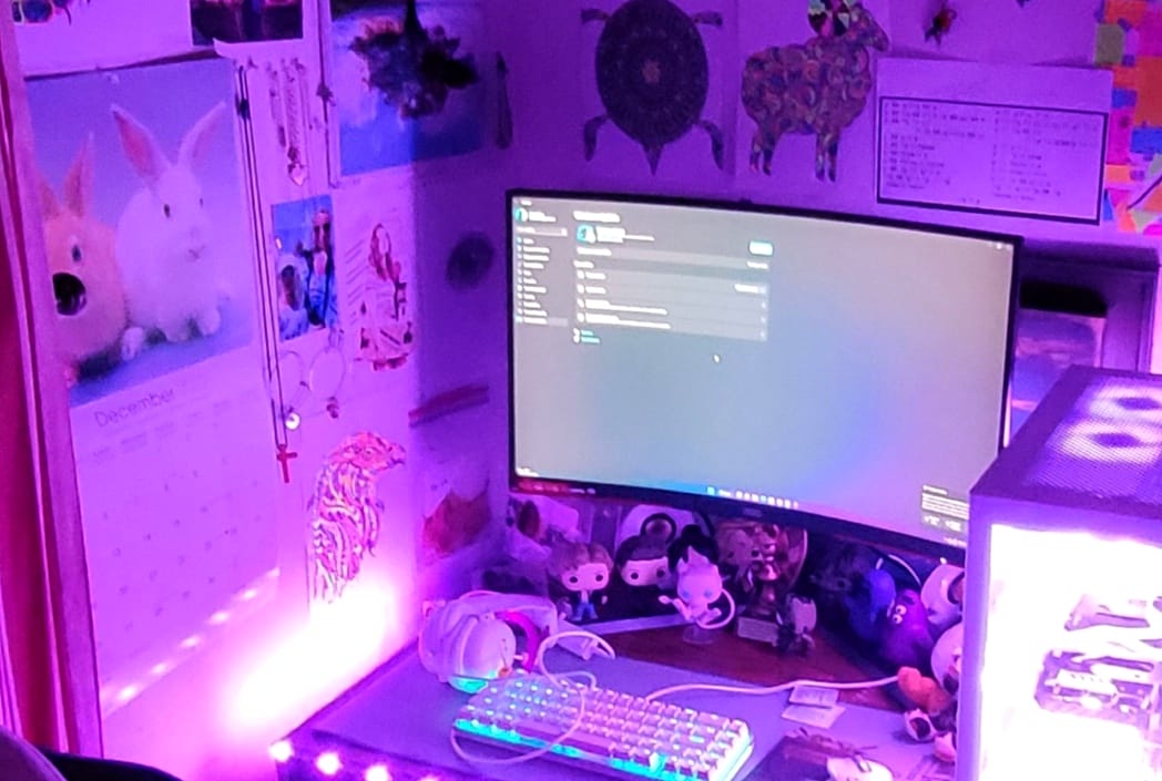 Una configuración de escritorio acogedora con iluminación púrpura con un monitor curvo que muestra la configuración. La decoración incluye Funko Pops, arte mural, un calendario y un teclado iluminado por el arco iris.
