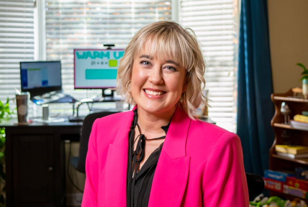 Una mujer sonriente con blazer rosa se para en la oficina en casa con una configuración de computadora borrosa en el fondo. Ambiente luminoso y profesional.