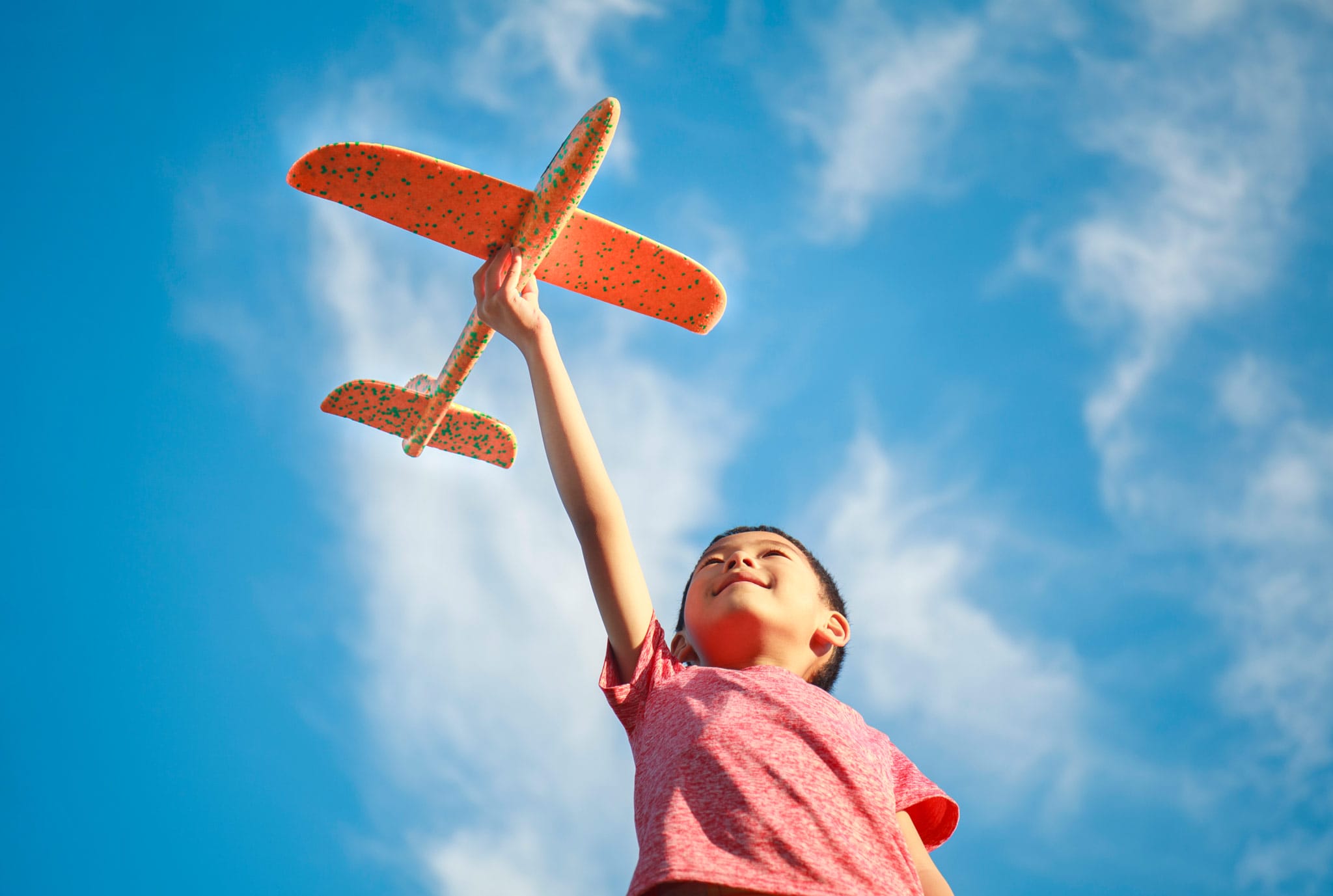 Un niño con una camisa roja sostiene un avión de juguete naranja en alto contra un cielo azul vibrante con nubes tenues, transmitiendo alegría e imaginación.