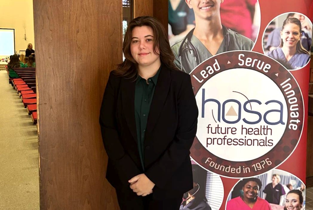 Joven con traje junto a un póster de HOSA en un pasillo. El cartel dice "Liderar, Servir, Innovar". El ambiente es formal y profesional.