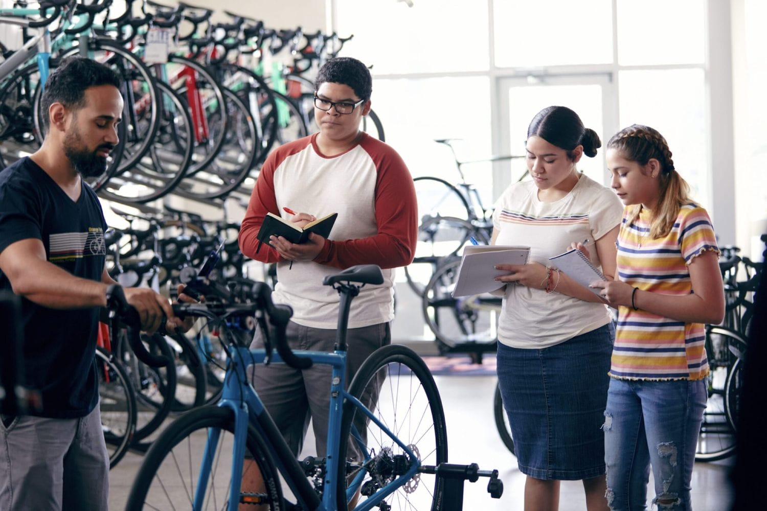 Hombre demostrando el montaje de bicicletas a tres estudiantes atentos tomando notas en una tienda de bicicletas bien iluminada. El fondo presenta filas de bicicletas coloridas.
