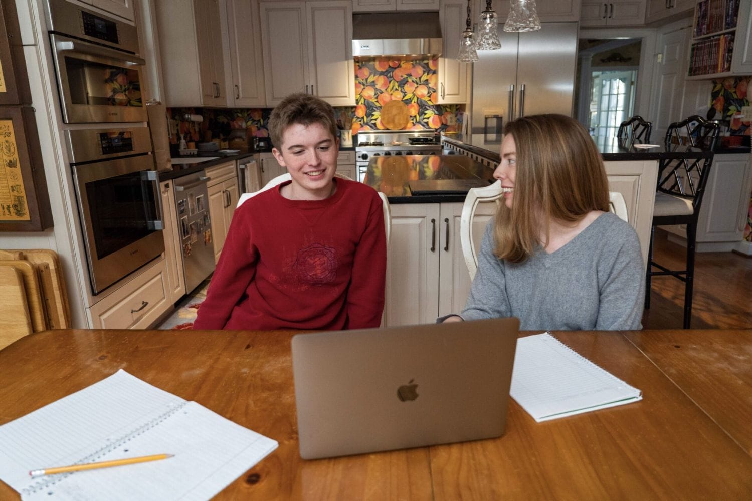 Two people sit at a kitchen table with a laptop and notebooks, smiling and engaged in conversation. The kitchen is modern with floral accents.
