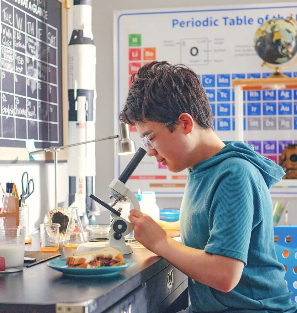 Niño examinando la comida bajo un microscopio en una habitación con temática científica, con una tabla periódica, una pizarra y un modelo de cohete, transmitiendo curiosidad.