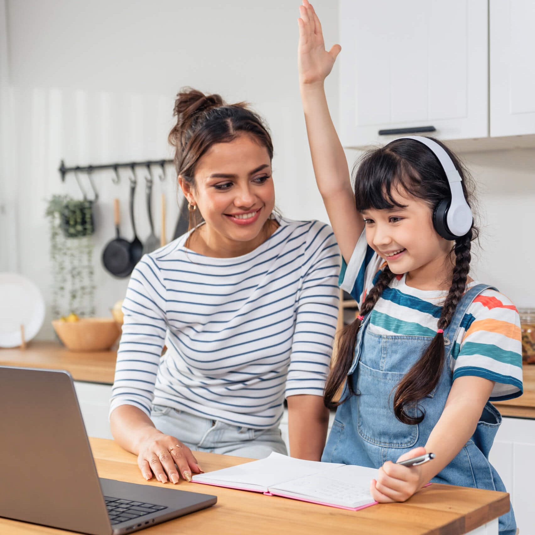 Una mujer sonriente con una camisa a rayas se sienta junto a una niña con auriculares y la mano levantada, trabajando en una mesa con una computadora portátil y un cuaderno en una cocina.