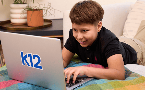 A smiling child lays on a colorful blanket, focused on a laptop displaying "k12." The scene is cozy and suggests engaged online learning.