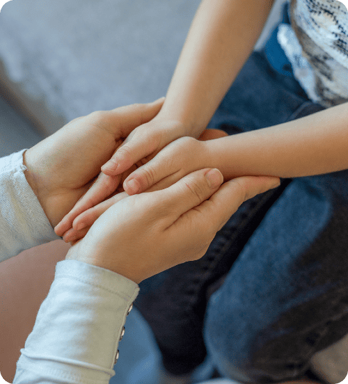 Hands of an adult gently holding a child's hands, conveying care and warmth. The child wears dark pants, and the tone suggests comfort and support.
