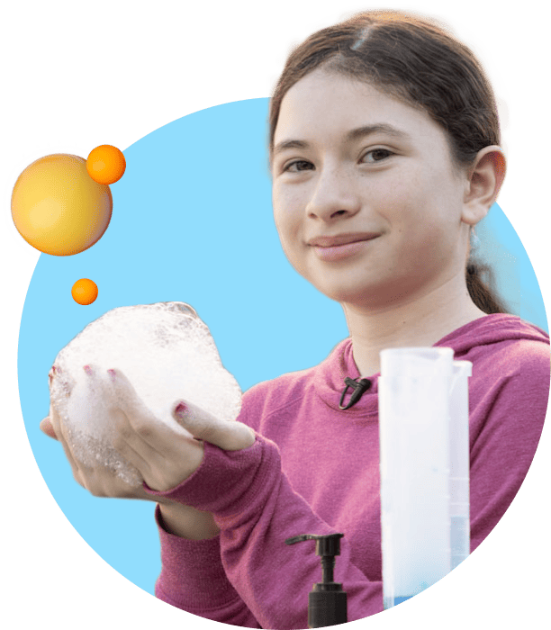 A young girl in a pink hoodie holds a large bubble foam in her hand, smiling against a light blue background with orange bubbles and a soap dispenser.