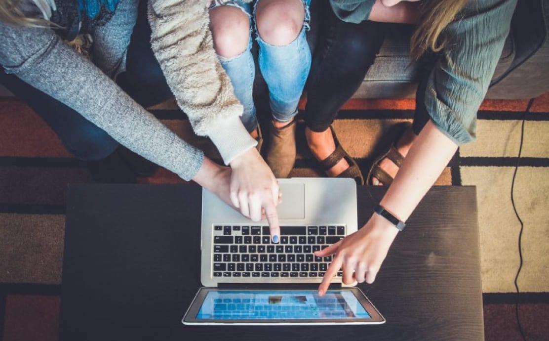Three people sit on a couch, leaning over a laptop on a coffee table. They point at the screen, suggesting collaboration and focus.
