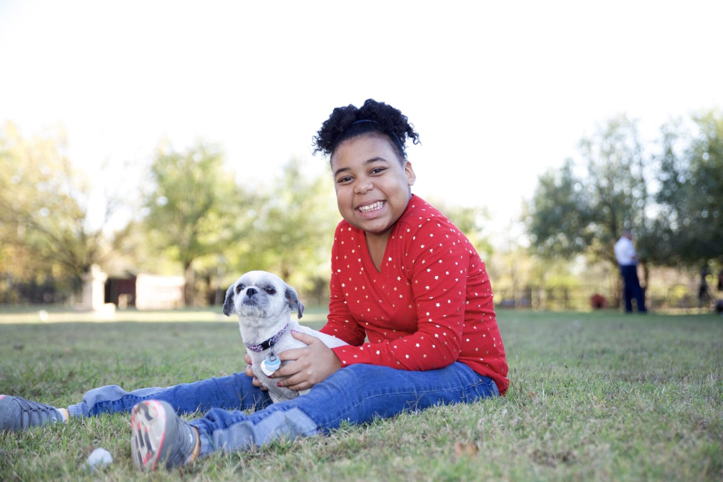 A child in a red sweater with white stars smiles while sitting on grass, holding a small dog. Trees and a person in the background suggest a park setting.