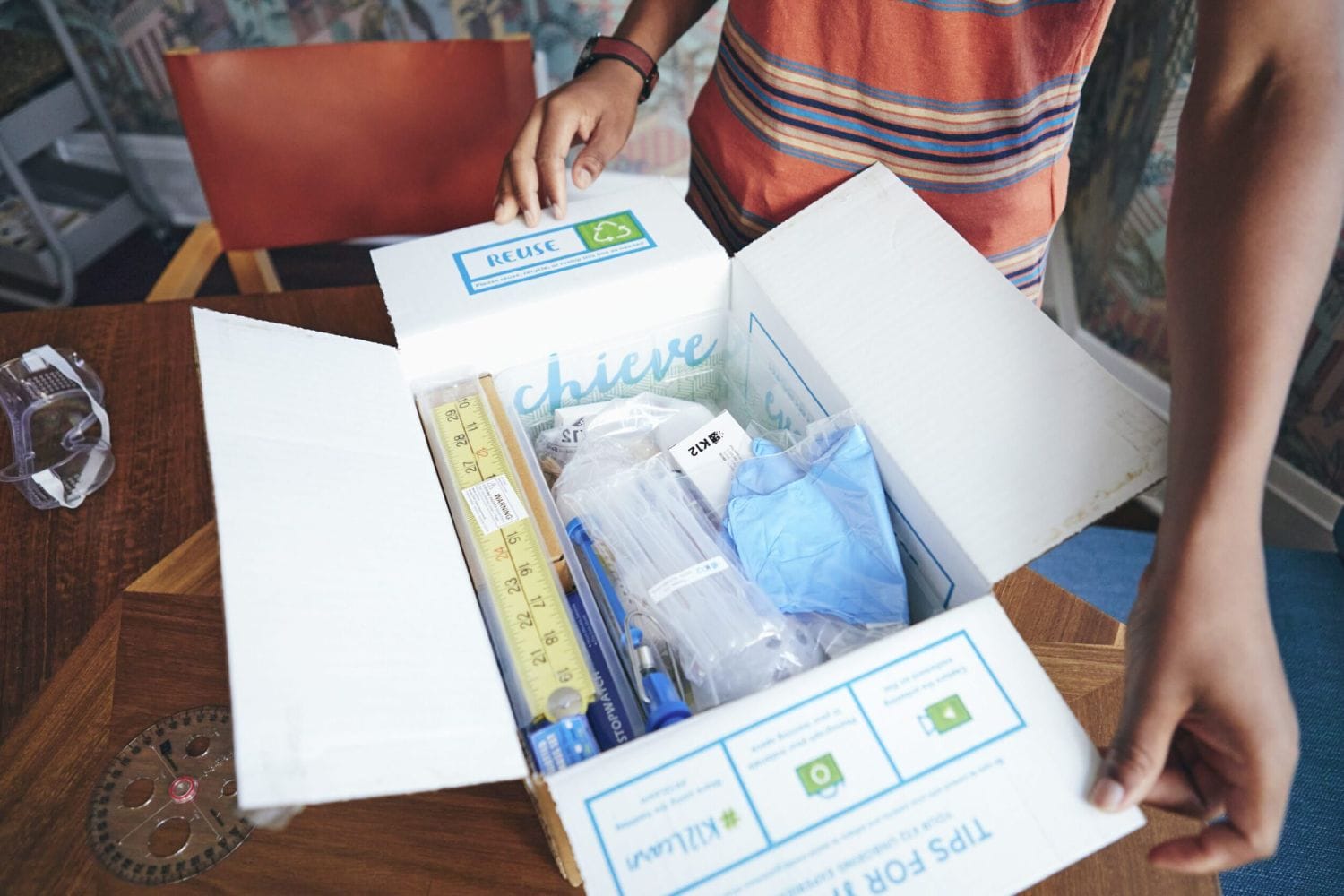 Una persona abre una caja con la etiqueta "REUTILIZAR" que contiene suministros educativos como reglas, guantes y bolígrafos, colocada en una mesa de madera con un transportador.