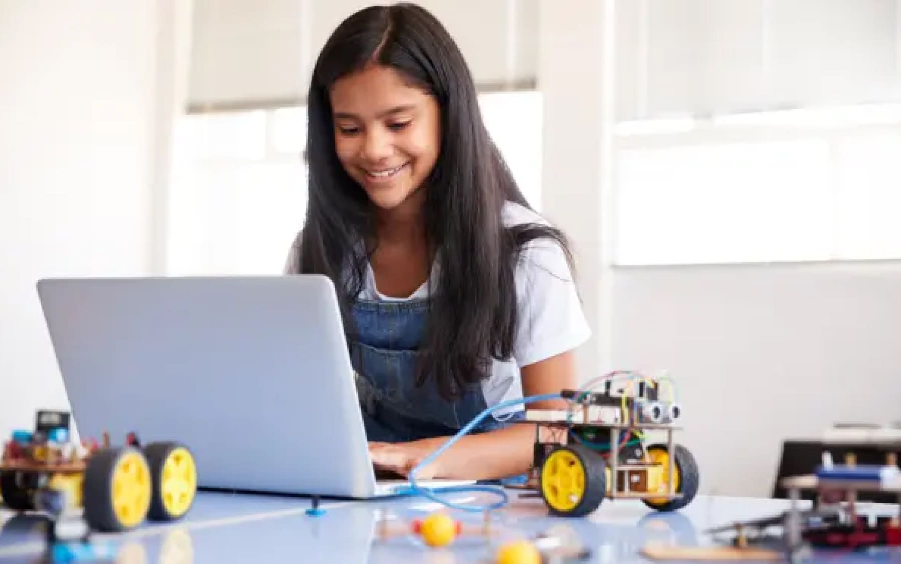 Una niña sonríe mientras trabaja en una computadora portátil en una mesa con proyectos de robótica. La habitación luminosa y soleada transmite una sensación de creatividad y aprendizaje.