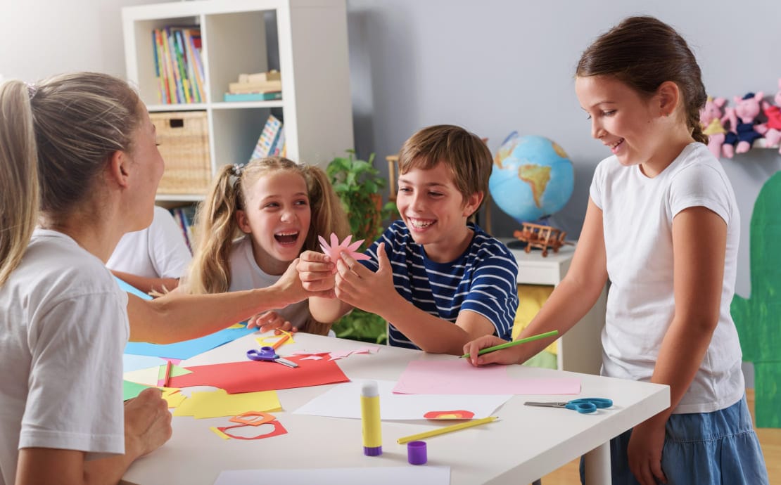 Los niños y un maestro están trabajando alegremente en un proyecto de manualidades en una mesa. La habitación es luminosa y colorida, con suministros de arte, un globo terráqueo y juguetes en el fondo.