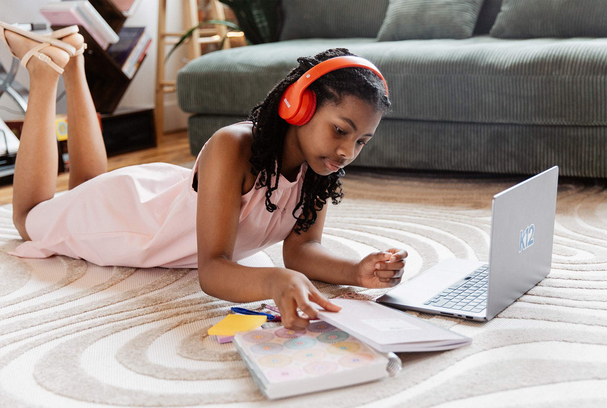 A young girl in pink dress lies on a carpet, wearing red headphones, studying with a laptop and notebook. The setting is a cozy living room.