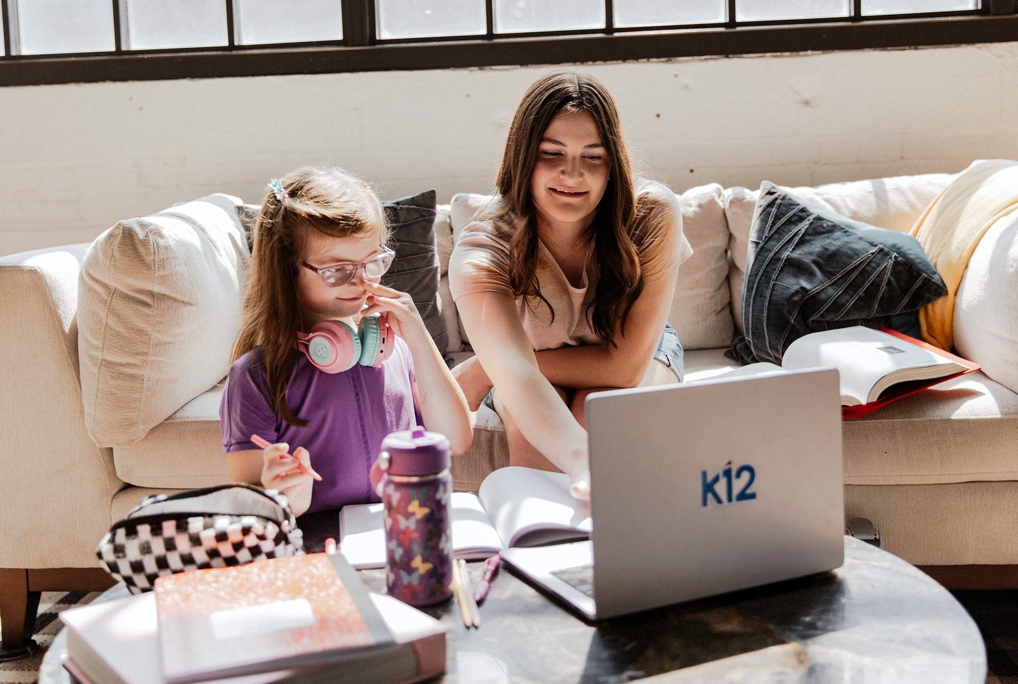Two girls sitting on a couch with a laptop labeled "k12." One is wearing headphones, and they appear focused and engaged in learning.