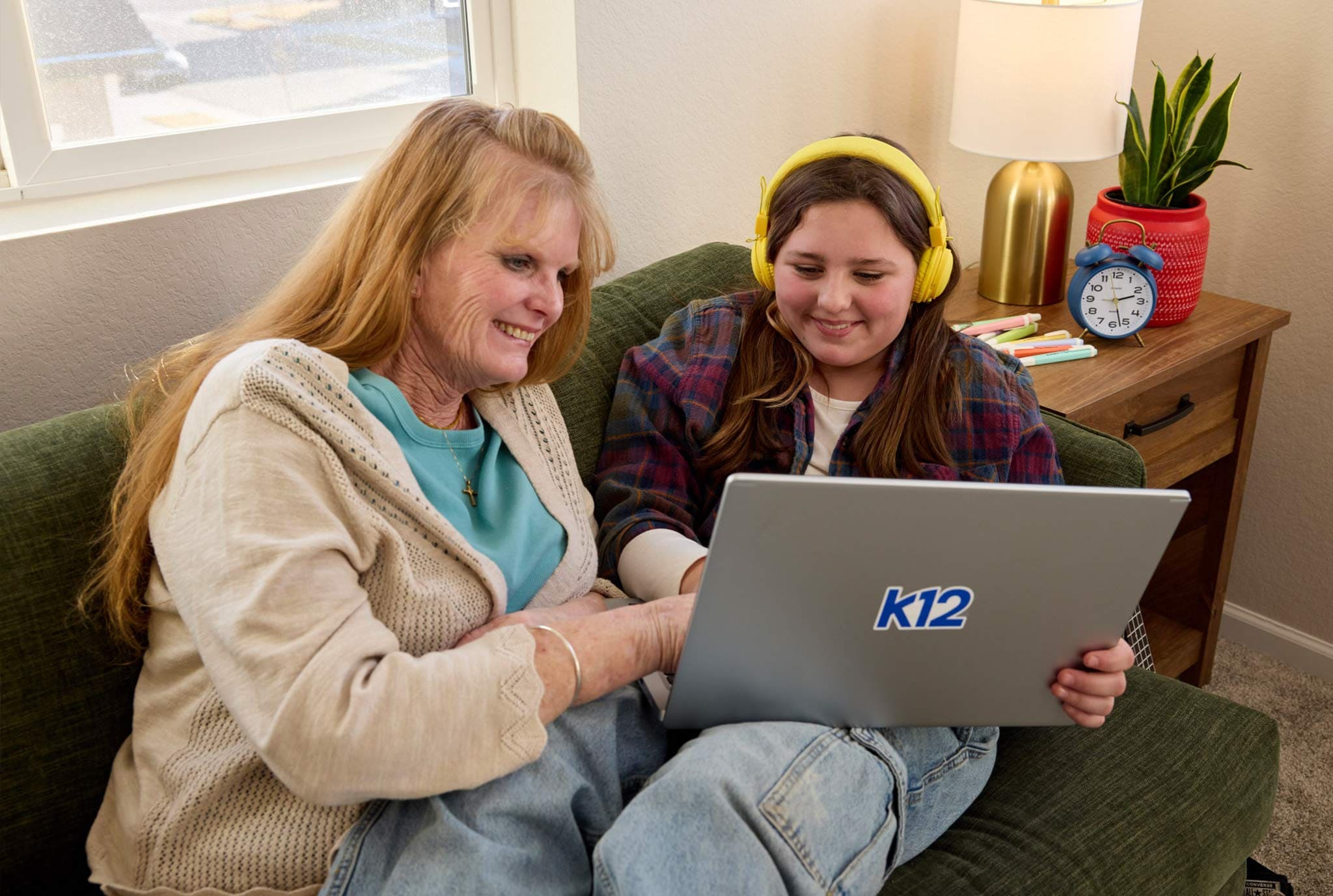 A woman and a young girl wearing yellow headphones sit on a couch, smiling at a K12 laptop. A cozy, inviting setting with a plant and clock nearby.