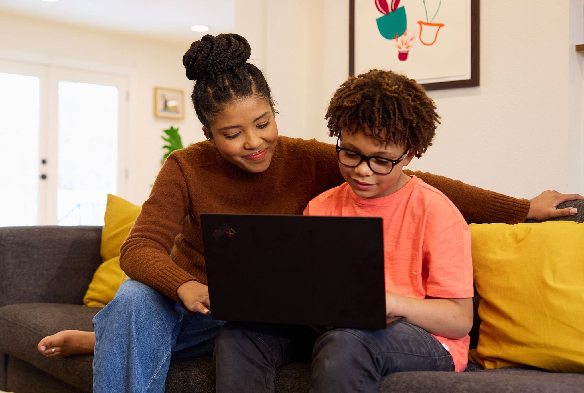 A woman and a boy sit closely on a couch, smiling while looking at a laptop. Bright cushions and a colorful wall art add warmth to the cozy setting.