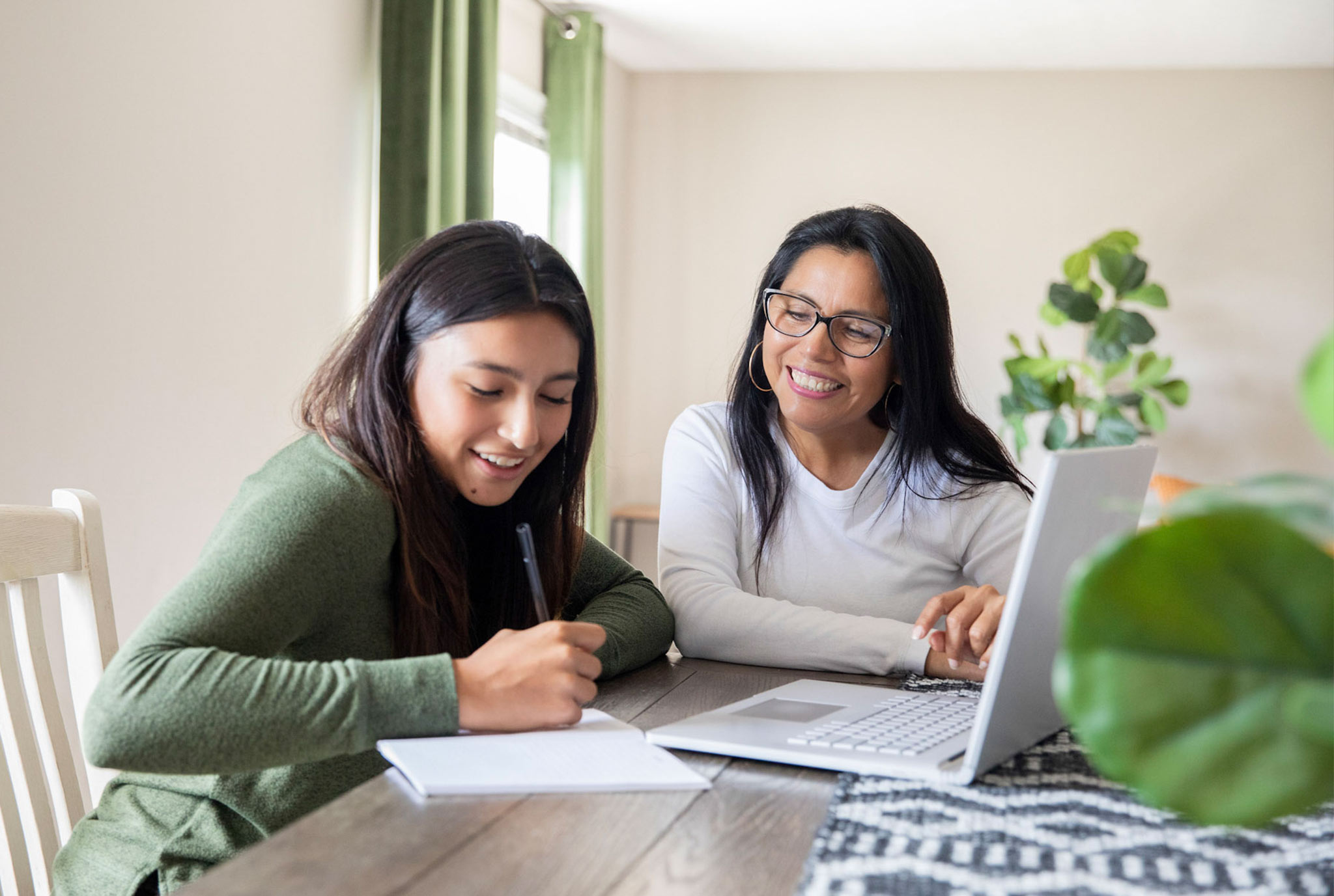 A woman and a young girl are seated at a table. The girl writes in a notebook while the woman smiles supportively. A laptop is open beside them.