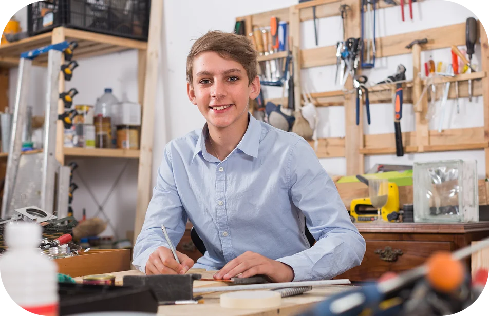 Teenage boy in a workshop smiles while sitting at a cluttered workbench, holding a pencil. Tools hang neatly on the wall, creating a creative vibe.