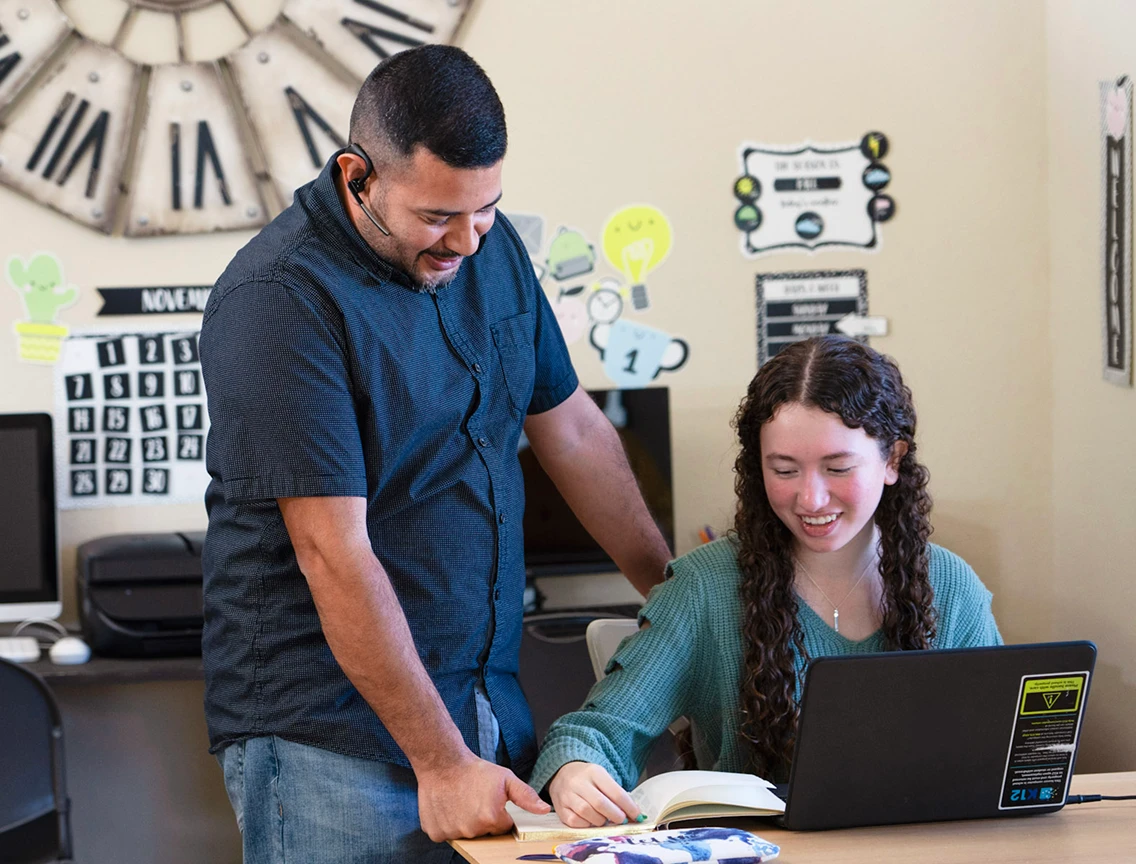 A teacher smiling and assisting a student in a classroom. The student is looking at a laptop, with a book open. The mood is supportive and collaborative.