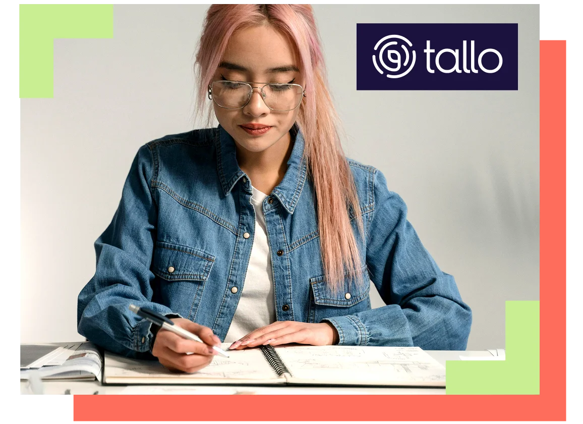 A person with pink hair and glasses writes in a notebook, focused and thoughtful. They're wearing a denim jacket. "Tallo" logo is in the top right corner.