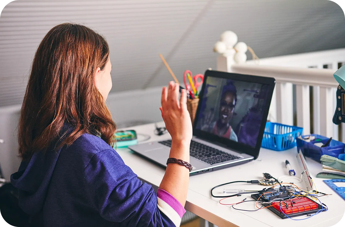 Una persona con ondas de cabello castaño en una computadora portátil durante una videollamada. Los componentes electrónicos se extienden sobre el escritorio. El entorno es informal y centrado en la tecnología.