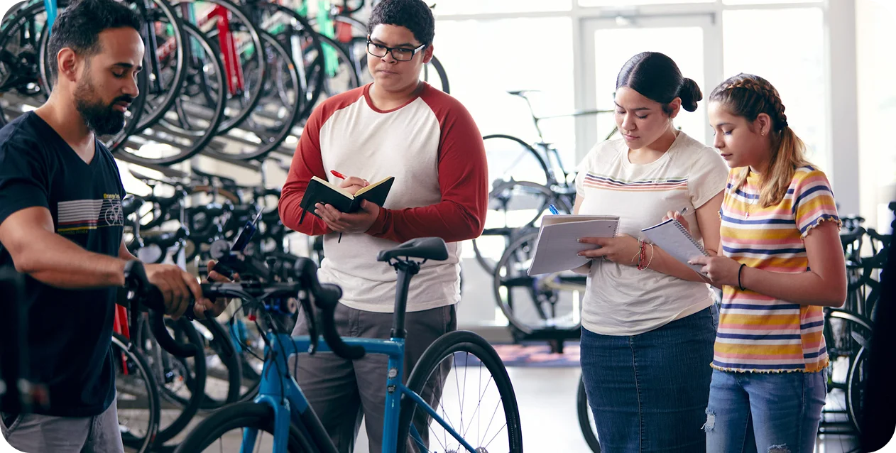 Un mecánico de bicicletas explica una bicicleta a tres estudiantes atentos con cuadernos en una tienda. La escena transmite una atmósfera de aprendizaje y concentración.