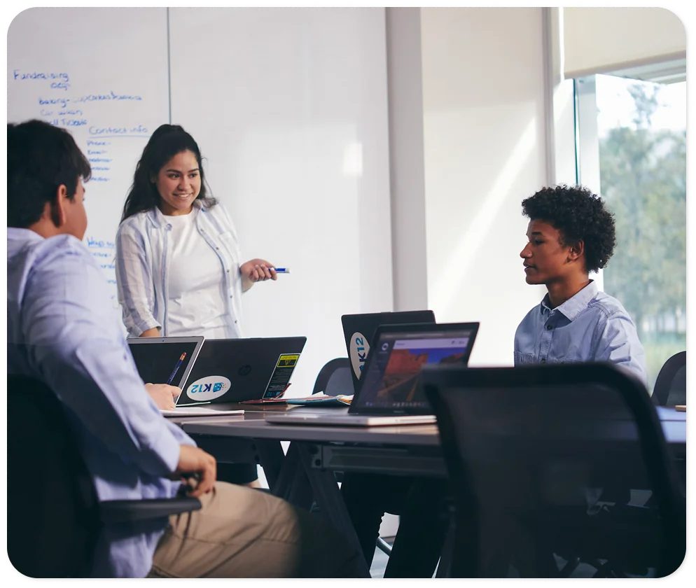 Un grupo diverso de adultos jóvenes colabora en una sala de reuniones luminosa, comprometida y atenta. Una mujer está de pie junto a una pizarra, sonriendo y sosteniendo un marcador, mientras que las computadoras portátiles están abiertas sobre una mesa.