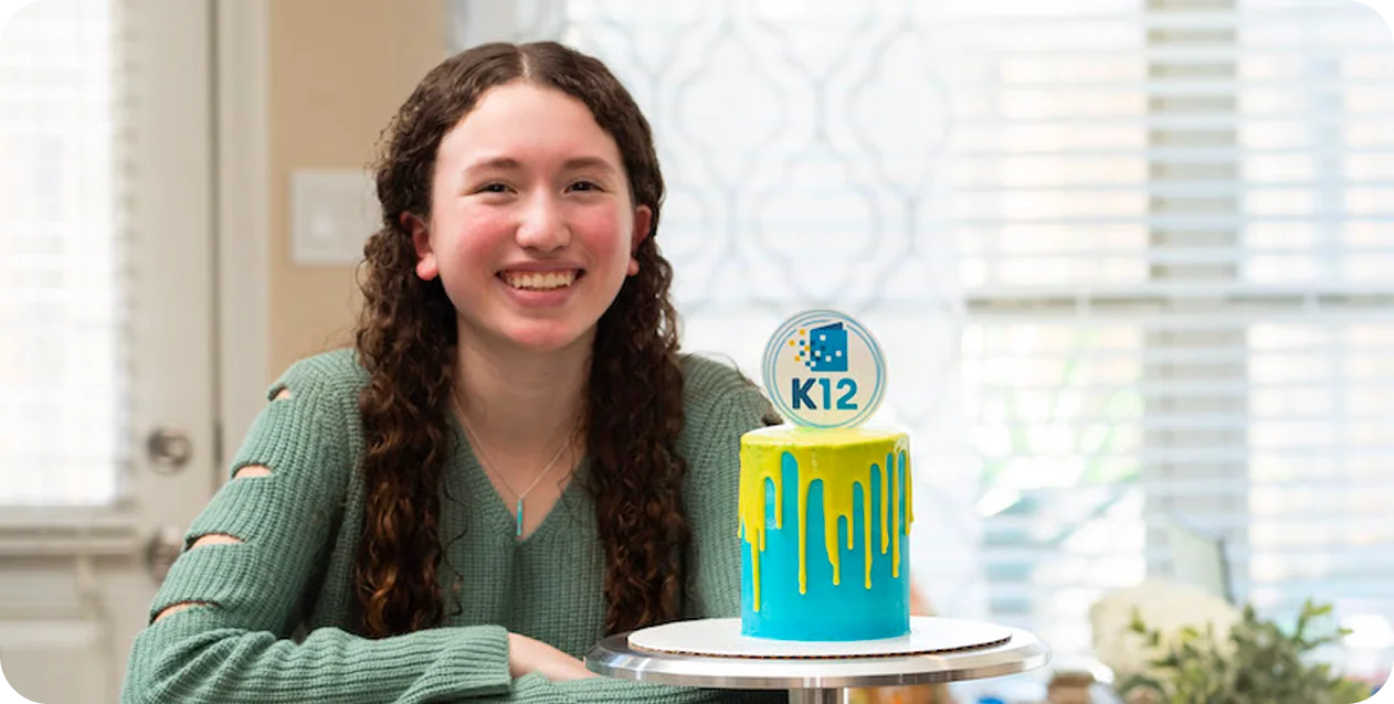Smiling girl in a green sweater sits beside a colorful cake with blue and yellow icing drips, topped with a "K12" sign. Bright, cheerful atmosphere.