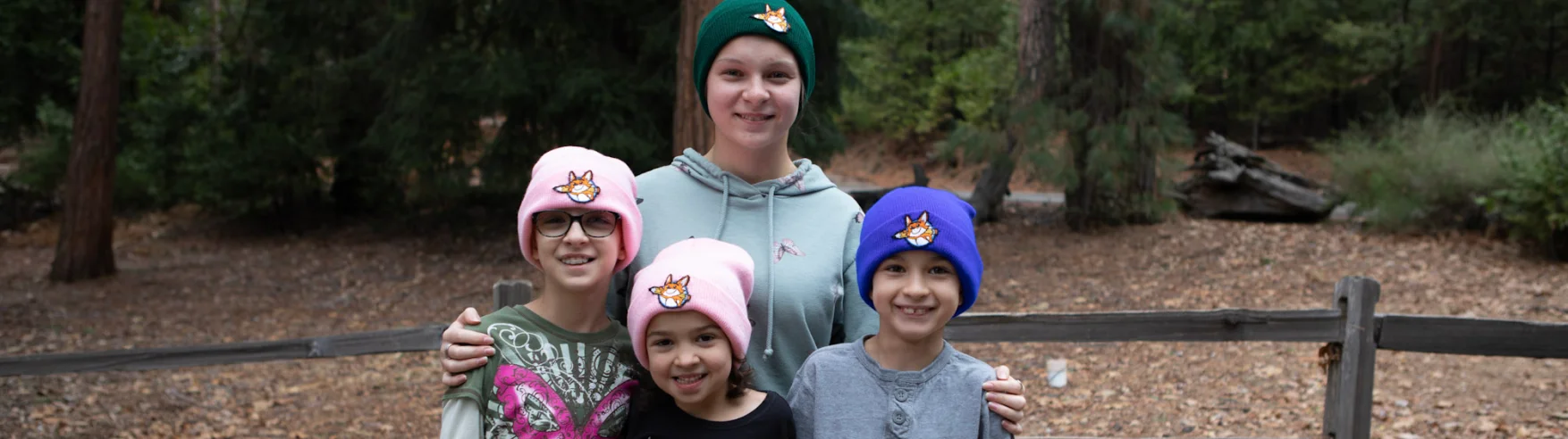 Un grupo de cuatro niños sonrientes se paran al aire libre en un bosque. Llevan gorros de colores con diseños de animales. El estado de ánimo es alegre y relajado.