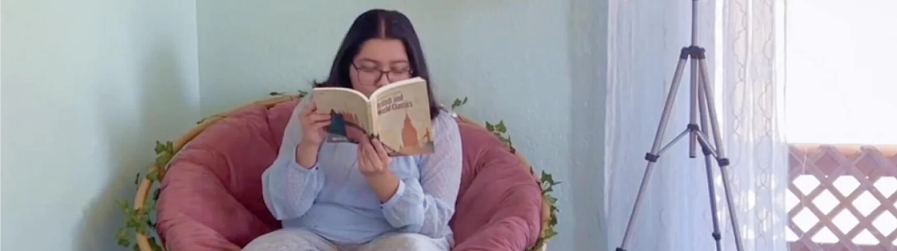 Una persona con cabello largo y oscuro se sienta en una silla grande y acolchada, leyendo un libro titulado "El Principito". Aparecen relajados en una habitación acogedora y suavemente iluminada.
