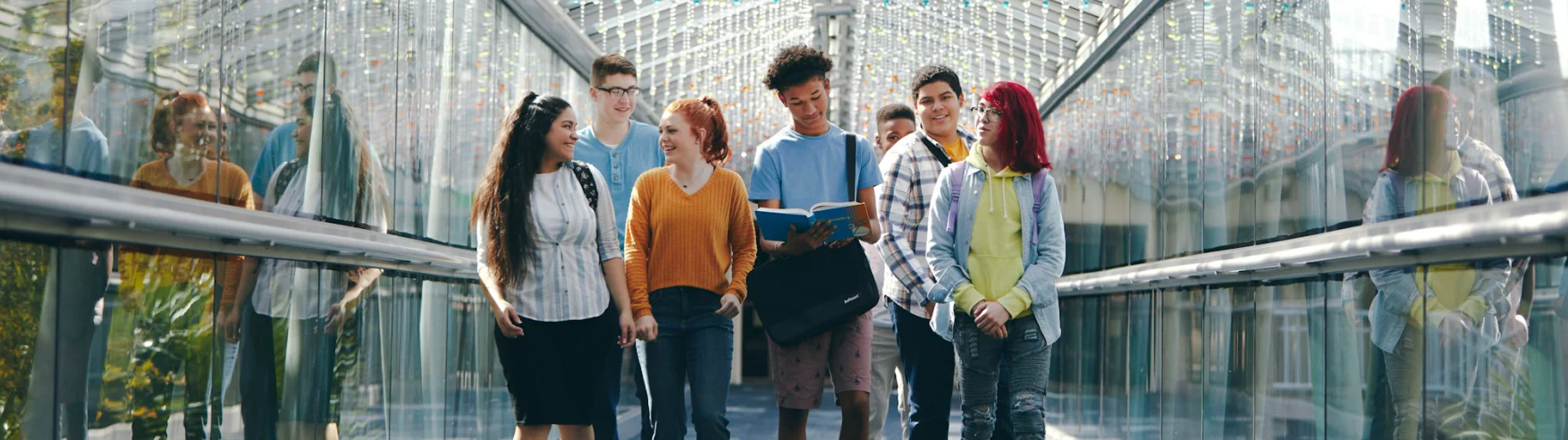 Un grupo diverso de seis estudiantes caminan por un pasillo de vidrio, sonriendo y charlando. Están vestidos de manera informal, creando un ambiente vibrante y juvenil.