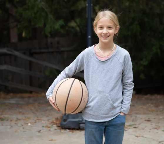 Una niña está parada afuera, sosteniendo una pelota de baloncesto debajo de un brazo. Lleva una sudadera con capucha gris y jeans, sonriendo contra un fondo de árboles y una cerca de madera.