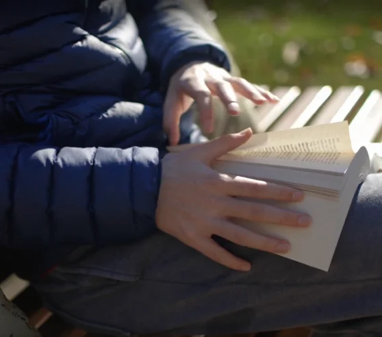 A person in a blue jacket is reading a book on a wooden bench in a sunny park. The scene conveys relaxation and a peaceful atmosphere.