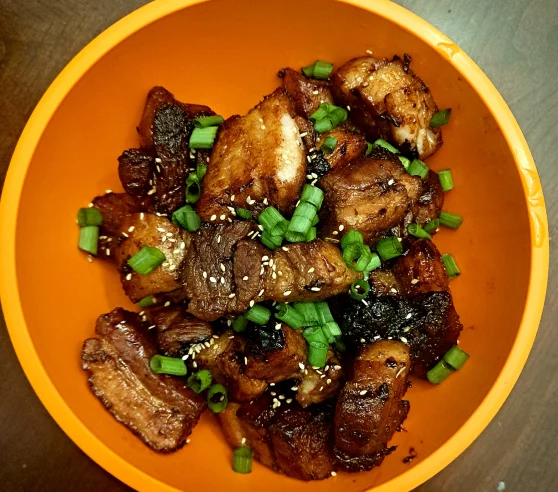 Un tazón naranja vibrante lleno de suculentos y crujientes trozos de panceta de cerdo adornados con semillas de sésamo y rodajas de cebolla verde fresca.