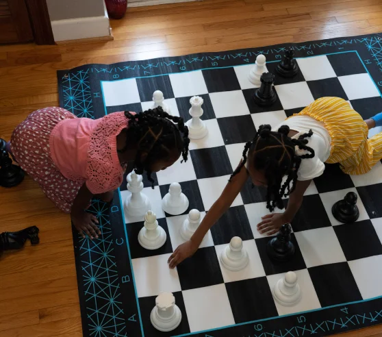 Two children play chess on a large floor mat. One in a pink top and skirt, the other in a white shirt and yellow skirt. They're focused and engaged.