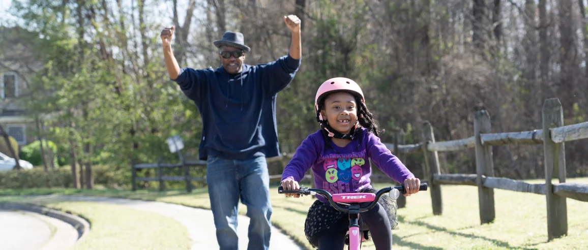 A joyful girl rides a pink bike on a sunny path, wearing a helmet and a purple shirt. Behind her, a man with raised arms cheers, expressing pride.