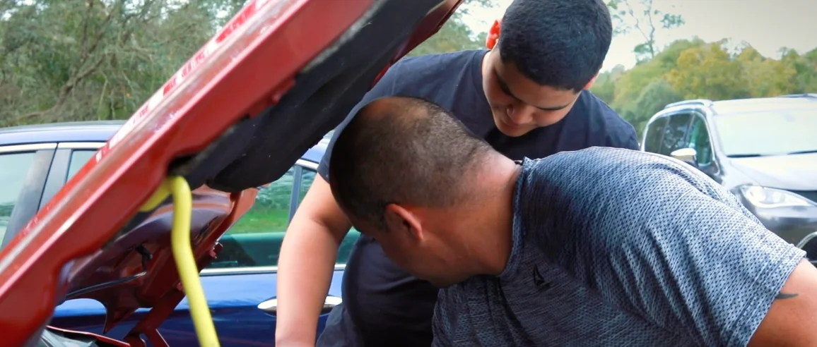 Dos personas están mirando debajo del capó de un automóvil, enfocadas y trabajando juntas. El entorno es al aire libre con otros coches y árboles en el fondo.
