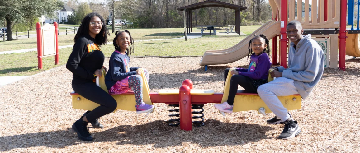 A family of four enjoying a sunny day at the park, sitting on a colorful seesaw. Two adults and two children, all smiling. Playground and trees in the background.