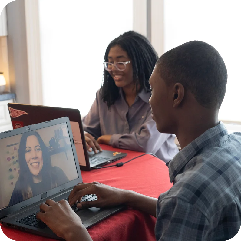 Two people work on laptops at a table with a red tablecloth. The person on the left smiles while on a video call, creating a positive, collaborative mood.